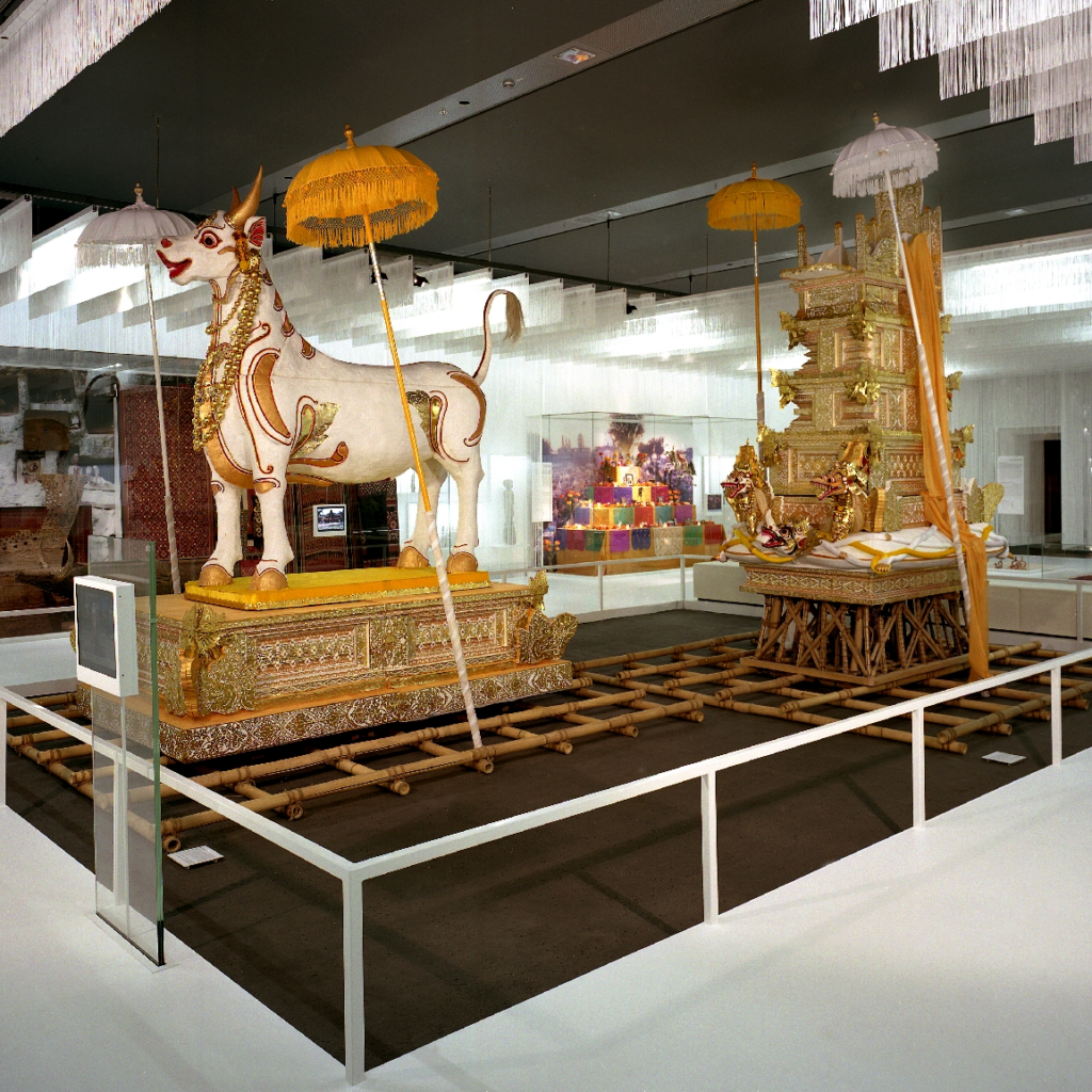 Ausstellungsraum im Rautenstrauch-Joest-Museum mit einer kunstvoll gestalteten Totenprozession: Im Vordergrund steht eine reich verzierte weiße Stierfigur auf einer vergoldeten Trageplattform, begleitet von zeremoniellen Schirmen. Dahinter befindet sich ein aufwendig geschmückter Leichen- oder Schreinaufbau auf Bambusgestellen. Die Installation thematisiert Abschied, Tod und Jenseits im Rahmen des Themenparcours „Der Mensch in seinen Welten“.