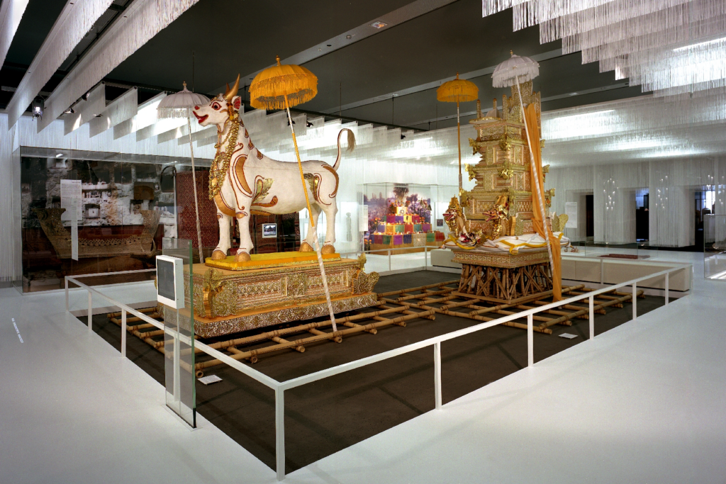 Ausstellungsraum im Rautenstrauch-Joest-Museum mit einer kunstvoll gestalteten Totenprozession: Im Vordergrund steht eine reich verzierte weiße Stierfigur auf einer vergoldeten Trageplattform, begleitet von zeremoniellen Schirmen. Dahinter befindet sich ein aufwendig geschmückter Leichen- oder Schreinaufbau auf Bambusgestellen. Die Installation thematisiert Abschied, Tod und Jenseits im Rahmen des Themenparcours „Der Mensch in seinen Welten“.