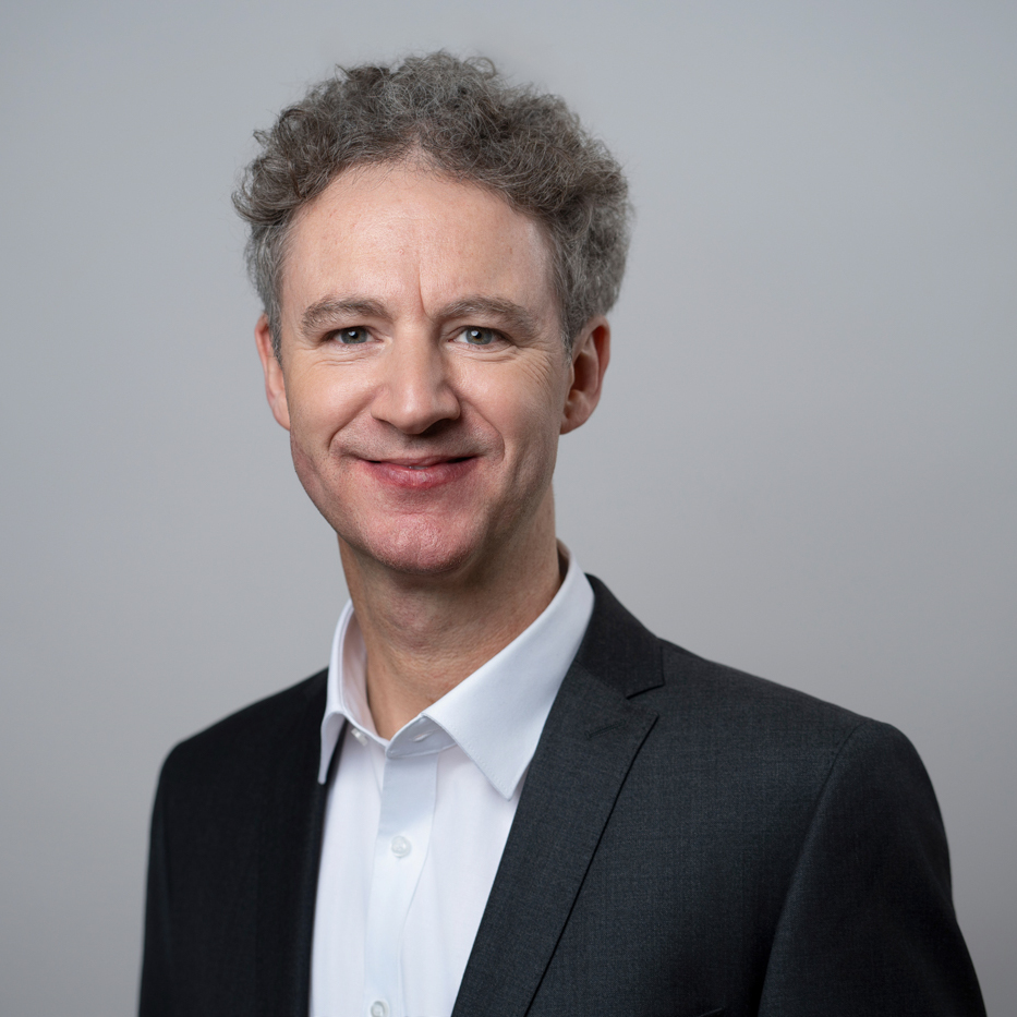 Porträt von Alexander Klein, einem Mann mittleren Alters mit kurzen, grauen Locken. Er lächelt und trägt einen schwarzes Jacket über einem weißen Hemd. Er steht vor einem grauen Hintergrund.