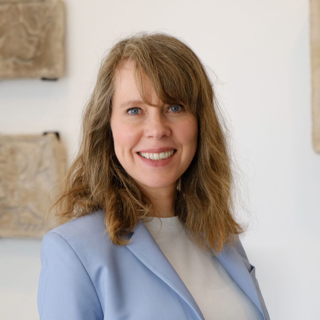 rträt von Barbara Foerster, einer Frau mittleren Alters mit schulterlangem, hellbraunem Haar. Sie lächelt und trägt einen hellblauen Blazer über einem weißen Oberteil. Sie steht vor einer hellen Wand mit dezenten Wandobjekten im Hintergrund.