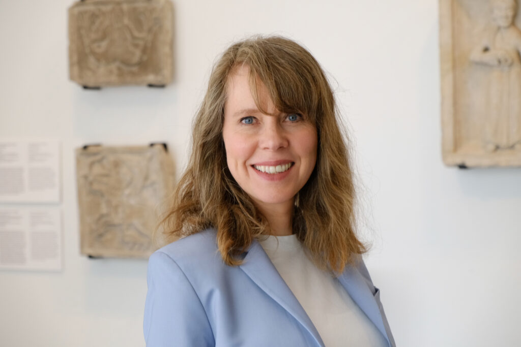 Porträt von Barbara Foerster, einer Frau mittleren Alters mit schulterlangem, hellbraunem Haar. Sie lächelt und trägt einen hellblauen Blazer über einem weißen Oberteil. Sie steht vor einer hellen Wand mit dezenten Wandobjekten im Hintergrund.