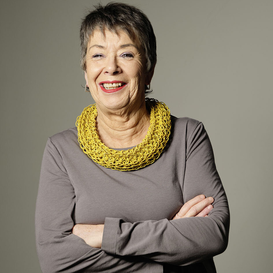 Porträt von Prof. Dr. Barbara Schock-Werner, einer älteren, lächelnden Frau mit kurzen grauen Haaren, die mit verschränkten Armen vor einem schlichten, hellen Hintergrund steht. Sie trägt ein langärmeliges graubraunes Kleid und eine auffällige, breite gelbe Halskette. Ihr freundlicher Gesichtsausdruck und roter Lippenstift vermitteln eine selbstbewusste, positive Ausstrahlung.