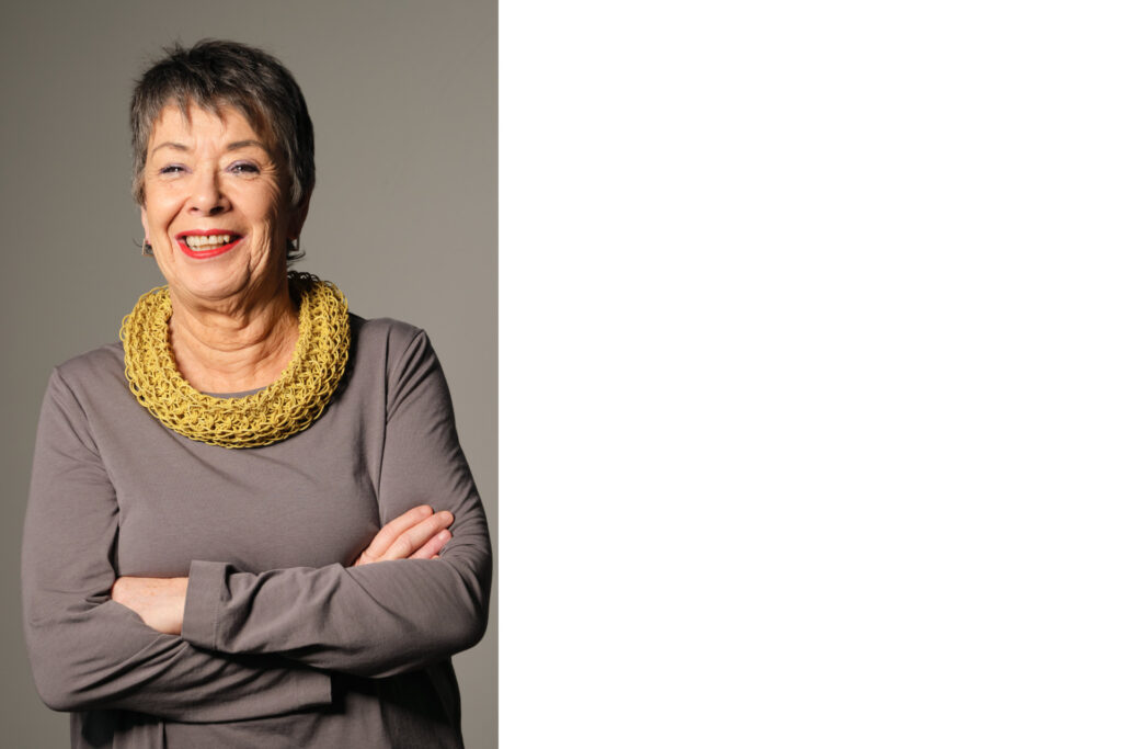Porträt von Prof. Dr. Barbara Schock-Werner, einer älteren, lächelnden Frau mit kurzen grauen Haaren, die mit verschränkten Armen vor einem schlichten, hellen Hintergrund steht. Sie trägt ein langärmeliges graubraunes Kleid und eine auffällige, breite gelbe Halskette. Ihr freundlicher Gesichtsausdruck und roter Lippenstift vermitteln eine selbstbewusste, positive Ausstrahlung.