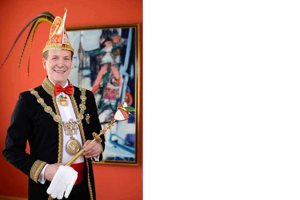 Christoph Kuckelkorn steht in seiner karnevalistischen Festkomitee-Uniform aus schwarzer Uniformjacke mit goldener und roter Spitze um den Rändern, weißem Hemd mit roter Fliege, roter Scherpe um den Bauch, Federhut sowie Karnevalsorden um den Hals. Er steht vor einer roten Wand mit einem abstrakten Gemälde, das ein Funkemariechen zeigt.