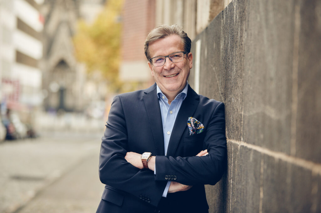 Porträt von Christoph Kuckelkorn, einem Mann mittleren Alters mit kurzem dunkelblondem und leicht grau meliertem Haar lehnt an einer Hauswand. Er trägt einen dunkelblauen Blazer und ein hellblaues Hemd. Er umschränkt die Arme und lächelt in die Kamera.