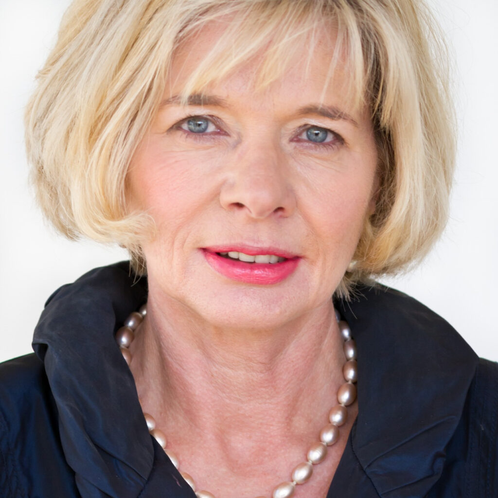 Porträtfoto von Cornelia C. Kliment, einer reifen Frau mit blondem, kinnlangem Haar, Perlenkette und schwarzer Bluse. Sie blickt lächelnd direkt in die Kamera.