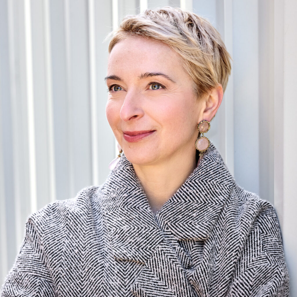 Ewa Bogusz-Moore, eine Frau mittleren Alters mit kurzem blonden Haar und in einem schwarz-weiß gemusterten Mantel, steht vor einer weißen Wand. Sie umschränkt die Arme und lächelt links an der Kamera vorbei.
