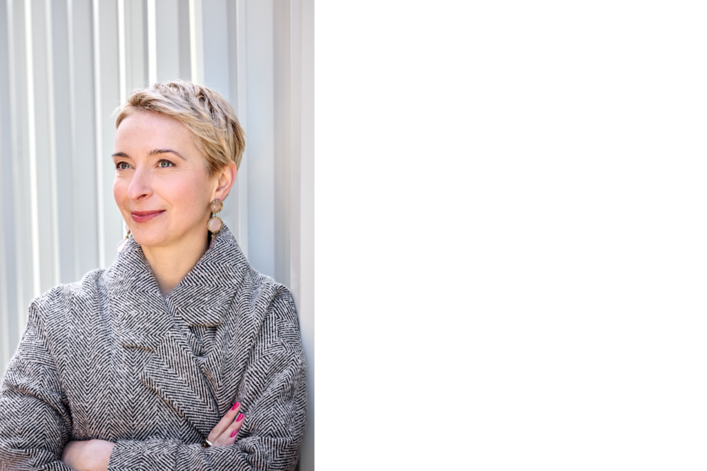 Ewa Bogusz-Moore, eine Frau mittleren Alters mit kurzem blonden Haar und in einem schwarz-weiß gemusterten Mantel, steht vor einer weißen Wand. Sie umschränkt die Arme und lächelt links an der Kamera vorbei.