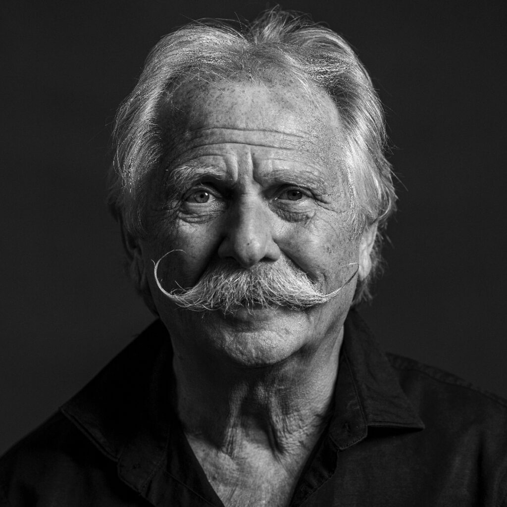 Ein schwarz-weiß-Porträt von Henning Krautmacher, einem älteren Mann mit vollem weißem Schnurrbart und zerzaustem Haar. Er trägt ein dunkles Hemd und blickt ruhig in die Kamera.