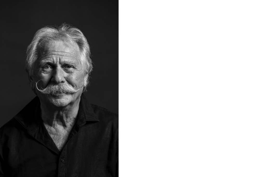 Ein schwarz-weiß-Porträt von Henning Krautmacher, einem älteren Mann mit vollem weißem Schnurrbart und zerzaustem Haar. Er trägt ein dunkles Hemd und blickt ruhig in die Kamera.