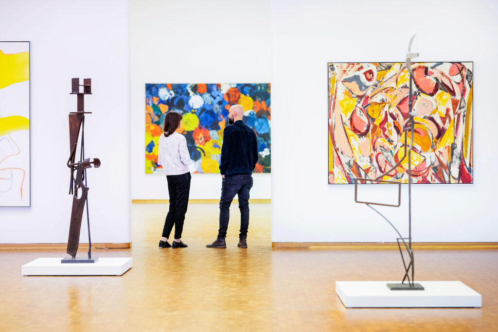 Heller Ausstellungsraum eines Kölner Museums mit Holzfußboden. Zwei Besucher*innen betrachten ein großformatiges, farbenfrohes abstraktes Gemälde. Im Raum stehen moderne Skulpturen auf weißen Sockeln, an den weißen Wänden hängen weitere abstrakte Kunstwerke. Die Szene zeigt eine Führung im Rahmen der Kulturvermittlung des Museumsdienstes Köln.