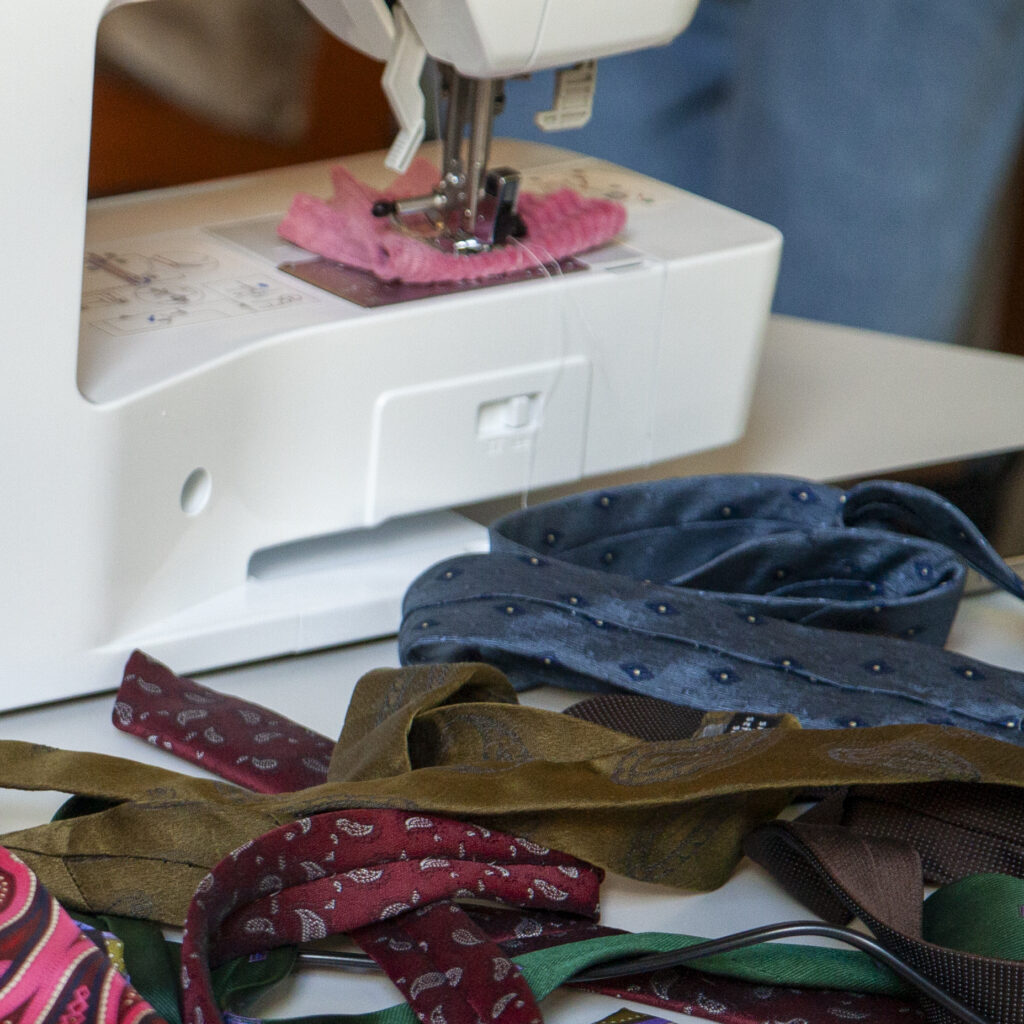 Eine weiße Nähmaschine der Marke Brother steht auf einem Tisch. Daneben liegen eine Schere, mehrere bunte Stoffstreifen und Krawatten. Die Nähmaschine ist eingeschaltet, ein Stück rosafarbener Stoff befindet sich unter dem Nähfuß. Die Szene wirkt wie ein Arbeitsplatz zum Nähen oder Reparieren von Textilien.