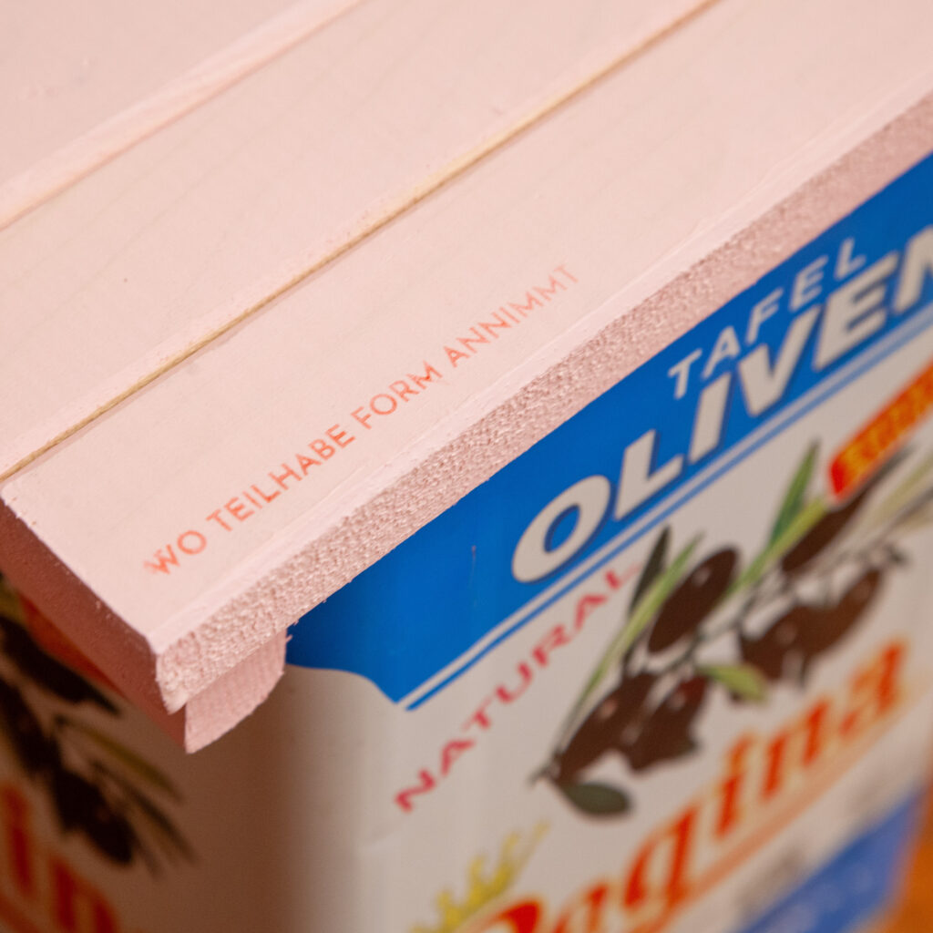 Nachhaltig gestalteter Beistelltisch aus Holz und einem recycelten Olivenölkanister, Detailansicht mit der Aufschrift „Wo Teilhabe Form annimmt“.