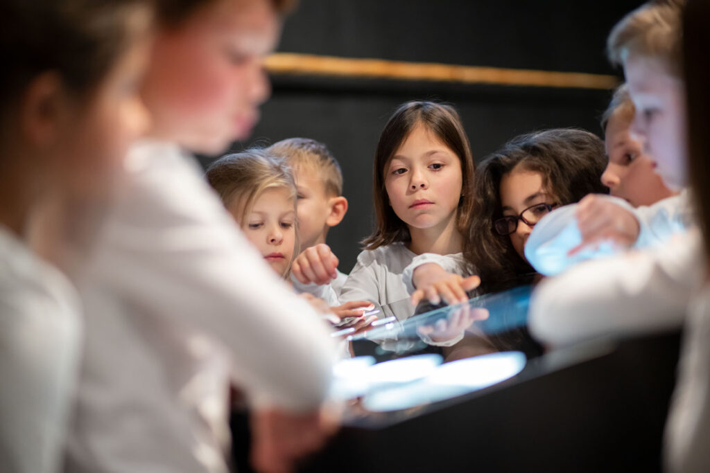Mehrere Kinder stehen um einen interaktiven Tisch in einem Museum und erkunden gemeinsam eine digitale Installation im Rahmen der Kulturvermittlung des Museumsdienstes Köln.