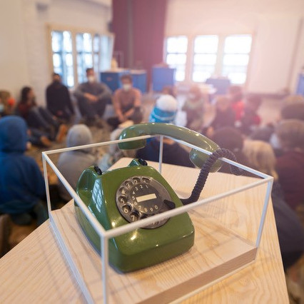 Ein grünes, altes Wählscheibentelefon steht in einer transparenten Plexiglasbox auf einem Holzpodest. Im unscharfen Hintergrund sitzen zahlreiche Menschen in einem hellen Raum im Kreis auf dem Boden, als Teil einer mobilen Ausstellung von Museen IN DER BOX.
