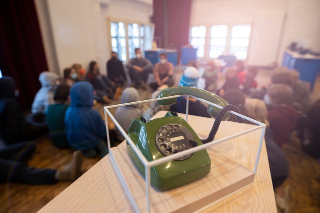 Ein grünes, altes Wählscheibentelefon steht in einer transparenten Plexiglasbox auf einem Holzpodest. Im unscharfen Hintergrund sitzen zahlreiche Menschen in einem hellen Raum im Kreis auf dem Boden, als Teil einer mobilen Ausstellung von Museen IN DER BOX.