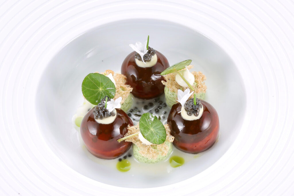 Ein perfekt angerichteter Teller mit Kaviertrüffeln. Die drei Kaviartrüffel sind im Dreieck auf einem weißen, tiefen Teller angerichtet und bestehen jeweils aus einer halbkugelförmigen braunen und hochglänzenden Gelee-Kuppel mit weißer, cremiger Haube und schwarzem Kaviar, geschmückt mit einer weißen Blüte und einem feinen grünen Keimling. Zwischen den Kaviartrüffeln sind je eine Halbkugel aus hellgrüner Kräutercreme mit einem luftig-knusprigen, beigefarbenen Chip bedeckt, auf dem ein kleiner weißer Cremetupfer und ein rundes, sattes Blatt platziert wurde. Alles ist auf einer weißen Soße mit schwarzen Kaviareiern angerichtet.