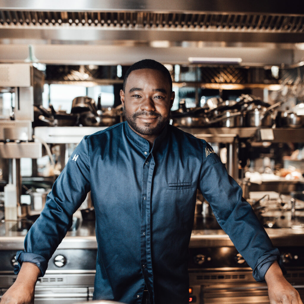 Porträt von Nelson Müller, einem jungen, dunkelhäutigen Mann mit kurzen schwarzen Haaren. Er trägt eine dunkelblaue Kochjacke und lächelt in die Kamera. steht in einer modernen Profiküche mit einem hohen Küchenregal aus Metall mit vielen Kochtöpfen im Hintergrund.