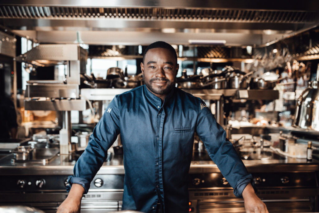 Porträt von Nelson Müller, einem jungen, dunkelhäutigen Mann mit kurzen schwarzen Haaren. Er trägt eine dunkelblaue Kochjacke und lächelt in die Kamera. steht in einer modernen Profiküche mit einem hohen Küchenregal aus Metall mit vielen Kochtöpfen im Hintergrund.
