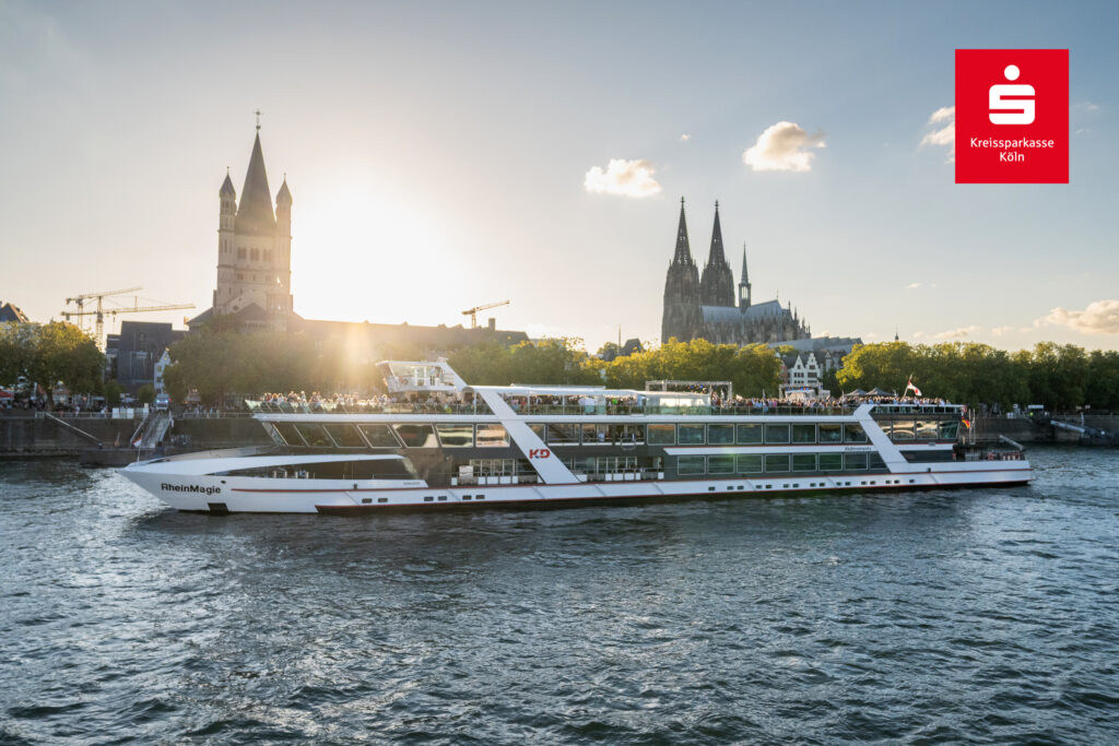 Ein Schiff mit dem Namen RheinMarie der KD Rheinschifffahrt fährt auf dem Rhein mit dem Kölner Dom rechts und der Kirche St. Severin links im Hintergrund. Die Sonne scheint und auf dem Schiffsdeck stehen und sitzen viele Menschen. Oben rechts das Logo der Kreissparkasse KölnBonn.