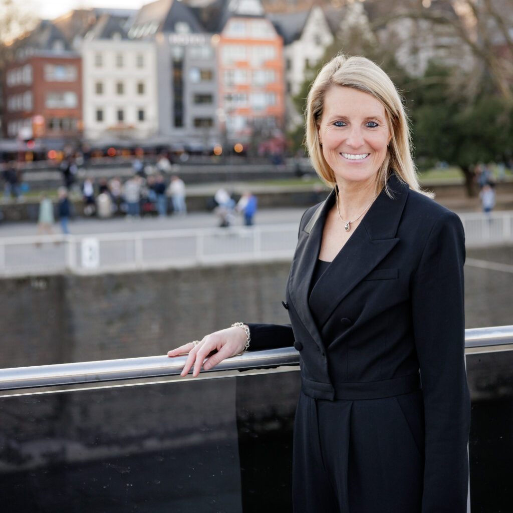 Porträt von Nina Luig, einer Frau mittleren Alters mit langem blondem Haar steht auf einem Schiff am Rheinufer in Köln, im Hintergrund ist der Kölner Dom und die Altstadt zu sehen. Sie trägt einen schwarzen Blazer und lächelt in die Kamera. Das Foto symbolisiert die Verbindung zwischen Rheinschifffahrt und Kultur in Köln.