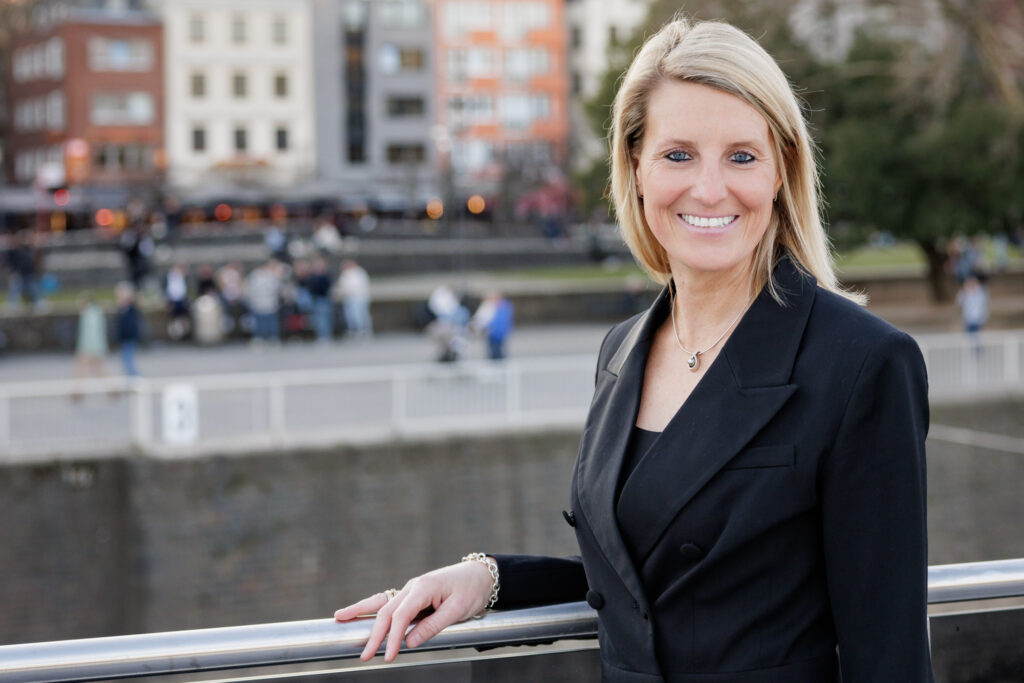 Porträt von Nina Luig, einer Frau mittleren Alters mit langem blondem Haar steht auf einem Schiff am Rheinufer in Köln, im Hintergrund ist der Kölner Dom und die Altstadt zu sehen. Sie trägt einen schwarzen Blazer und lächelt in die Kamera. Das Foto symbolisiert die Verbindung zwischen Rheinschifffahrt und Kultur in Köln.