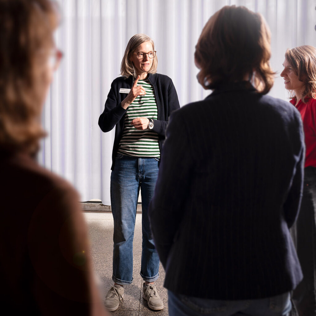 Eine Frau mit schulterlangen hellen Haaren und Brille steht im Mittelpunkt einer Gruppe und erklärt etwas mit einer Handbewegung. Sie trägt ein gestreiftes Shirt, eine dunkle Jacke und Jeans. Um sie herum sind mehrere Personen von hinten oder seitlich zu sehen, die ihr aufmerksam zuhören. Die Szene zeigt die Vermittlerin Frau Schwede bei einer Führung in Deutscher Gebärdensprache (DGS) im Museum für Angewandte Kunst Köln (MAKK).