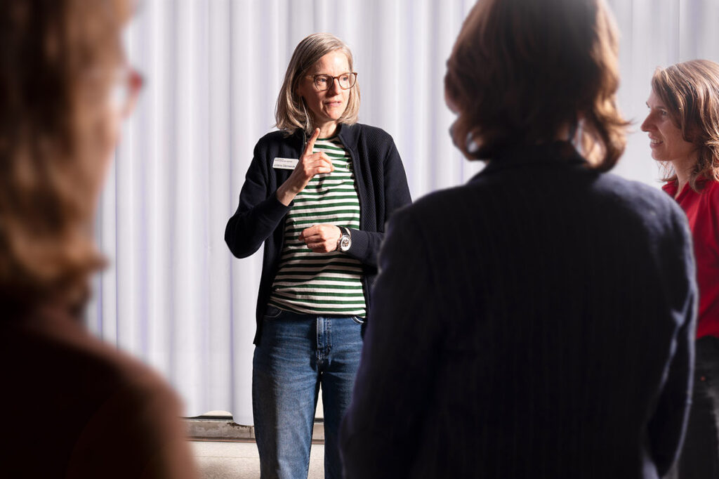Eine Frau mit schulterlangen hellen Haaren und Brille steht im Mittelpunkt einer Gruppe und erklärt etwas mit einer Handbewegung. Sie trägt ein gestreiftes Shirt, eine dunkle Jacke und Jeans. Um sie herum sind mehrere Personen von hinten oder seitlich zu sehen, die ihr aufmerksam zuhören. Die Szene zeigt die Vermittlerin Frau Schwede bei einer Führung in Deutscher Gebärdensprache (DGS) im Museum für Angewandte Kunst Köln (MAKK).