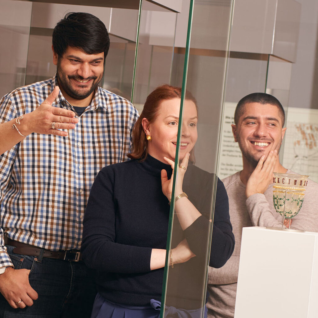 Drei Personen mit unterschiedlichen Herkünften stehen vor einem Kelch aus Glas in einem Glaskasten, das Ihnen gezeigt wird.