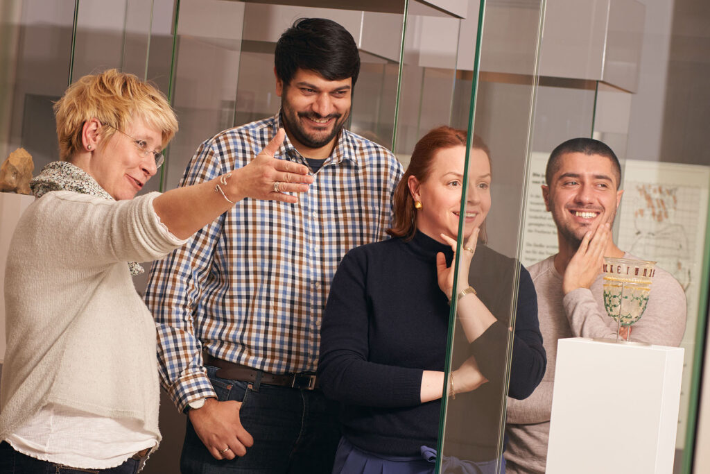 Drei Personen mit unterschiedlichen Herkünften stehen vor einem Kelch aus Glas in einem Glaskasten, das Ihnen gezeigt wird.