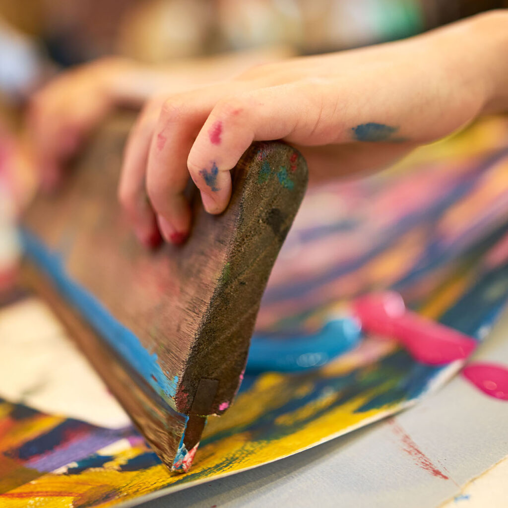 Eine mit Farbflecken bedeckte Kinderhand hält ein Holzrakel und verteilt bunte Farbe auf einem Blatt Papier; im Vordergrund sind leuchtende Blau-, Gelb- und Pinktöne zu sehen.
