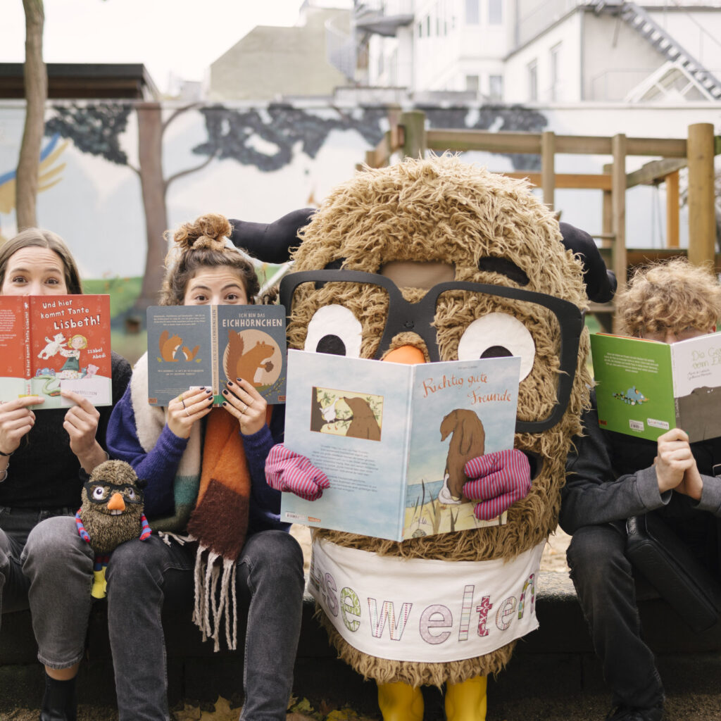 Auf dem Bild sieht man vier „Leser“ auf einer Parkbank, die Kinderbücher in den Händen halten und direkt in die Kamera schauen. Von links nach rechts: Eine Frau mit langen braunen Haaren, die ein rotes Kinderbuch mit dem Titel "Und hier kommt Tante Lisbeth!" hält. Eine zweite Frau mit hochgestecktem Haar und einem bunten Schal, die ein Buch über Eichhörnchen hält. Eine Person in einem großen, flauschigen Tierkostüm – offenbar eine Eule oder ein anderes Tier mit Brille – die ein Buch mit dem Titel "Richtig gute Freunde liest". Eine Person mit lockigem, hellbraunem Haar, die ein grünes Buch mit dem Titel "Die Geschichte vom Elefanten" hält. Die Szene spielt draußen auf einem Spielplatz, im Hintergrund ist eine Wand bemalt mit einer Landschaft und einem blauen Vogel. Auf dem Schoß der zweiten Person von links sitzt außerdem ein kleines Plüschtier, das wie die kostümierte Figur aussieht.