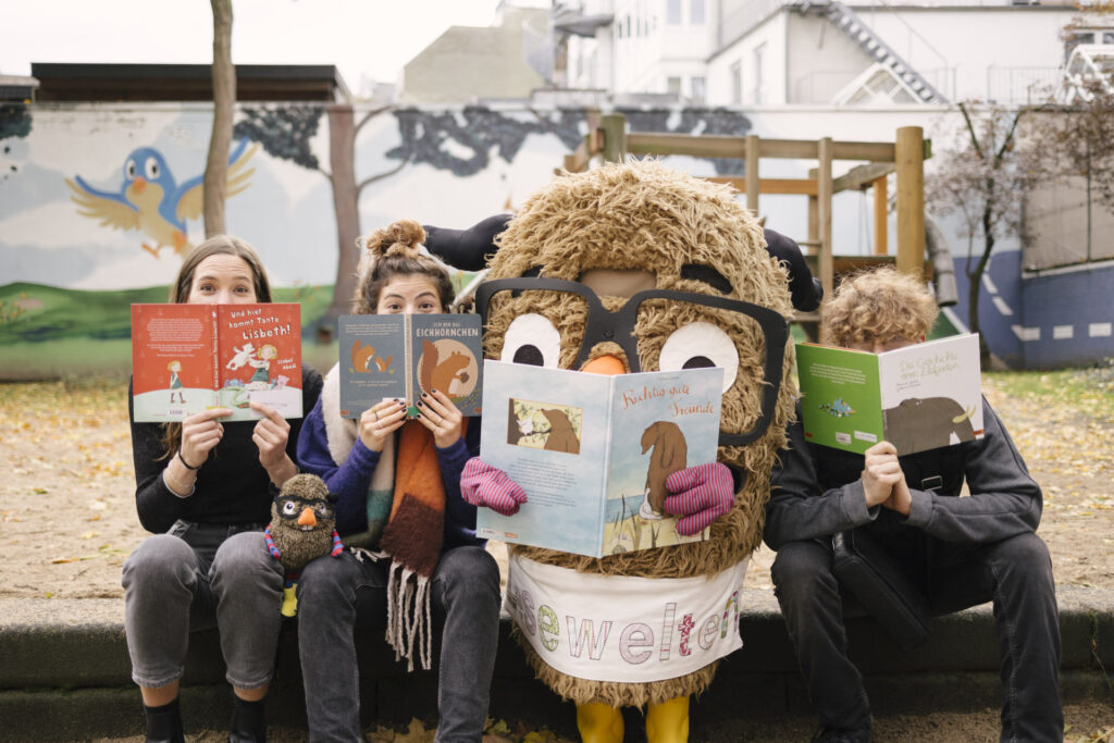 Auf dem Bild sieht man vier „Leser“ auf einer Parkbank, die Kinderbücher in den Händen halten und direkt in die Kamera schauen. Von links nach rechts: Eine Frau mit langen braunen Haaren, die ein rotes Kinderbuch mit dem Titel "Und hier kommt Tante Lisbeth!" hält. Eine zweite Frau mit hochgestecktem Haar und einem bunten Schal, die ein Buch über Eichhörnchen hält. Eine Person in einem großen, flauschigen Tierkostüm – offenbar eine Eule oder ein anderes Tier mit Brille – die ein Buch mit dem Titel "Richtig gute Freunde liest". Eine Person mit lockigem, hellbraunem Haar, die ein grünes Buch mit dem Titel "Die Geschichte vom Elefanten" hält. Die Szene spielt draußen auf einem Spielplatz, im Hintergrund ist eine Wand bemalt mit einer Landschaft und einem blauen Vogel. Auf dem Schoß der zweiten Person von links sitzt außerdem ein kleines Plüschtier, das wie die kostümierte Figur aussieht.